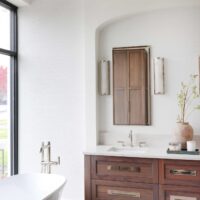The modern classic primary bath features walnut console vanity topped with quartzite countertop and paired with a textured wallpaper backsplash. Freestanding tub makes a sculptural statement on a porcelain tile floor.