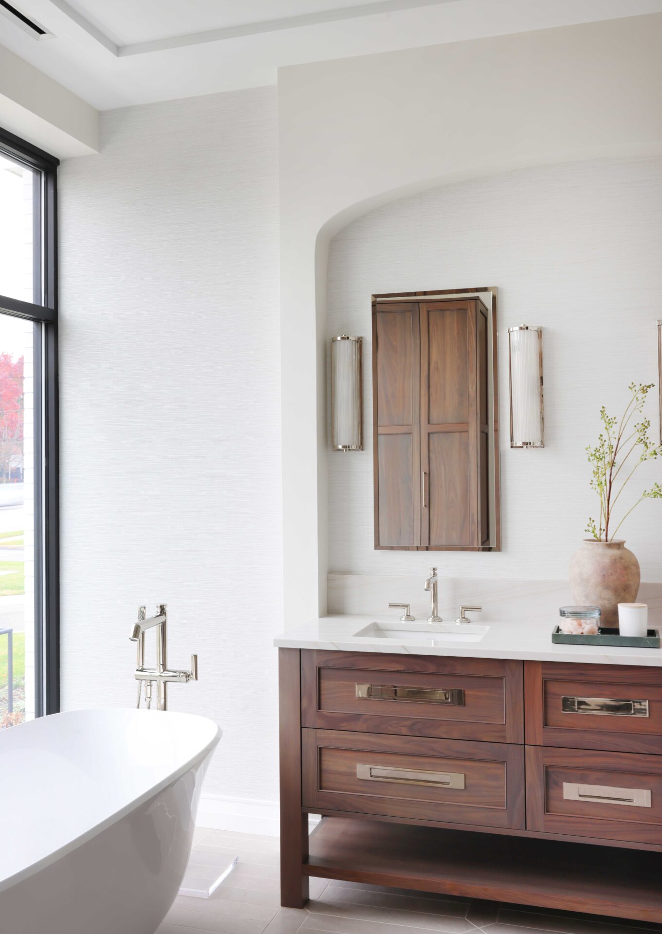 The modern classic primary bath features walnut console vanity topped with quartzite countertop and paired with a textured wallpaper backsplash. Freestanding tub makes a sculptural statement on a porcelain tile floor.