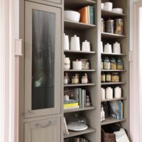 Garden estate butlers pantry features generous open shelving for everyday storage and display; custom cabinetry is painted in a soft, earthy mushroom tone with a brushed glaze and features vintage-style cutouts, furniture-like proportions, and tall baseboards that ground the space. Inside the cabinets are bottle holders, tip-outs for laundry or pet food, and thoughtful storage solutions tailored to daily life.