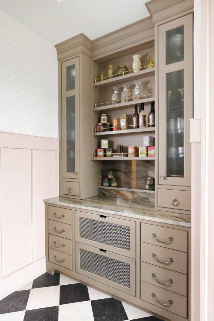 Garden estate butlers pantry features generous open shelving for everyday storage and display; custom cabinetry is painted in a soft, earthy mushroom tone with a brushed glaze and features vintage-style cutouts, furniture-like proportions, and tall baseboards that ground the space. Lattice mesh produce drawers — vented at both the front and bottom — bring in a distinctly garden-inspired detail, ideal for storing fruits and vegetables. Inside the cabinets are bottle holders, tip-outs for laundry or pet food, and thoughtful storage solutions tailored to daily life. An earthy-toned quartzite plays a starring role as well, wrapping the countertops, backsplash, and even the sculptural sink, all cut from the same slab for a seamless look.