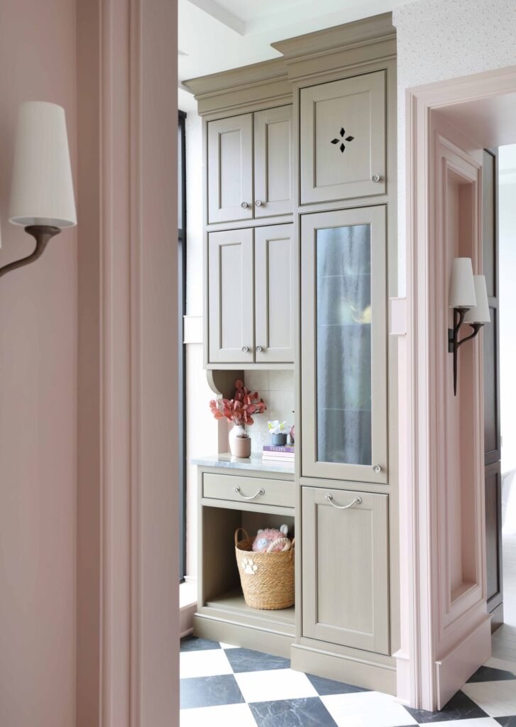 Garden estate butlers pantry features generous open shelving for everyday storage and display; custom cabinetry is painted in a soft, earthy mushroom tone with a brushed glaze and features vintage-style cutouts, furniture-like proportions, and tall baseboards that ground the space. Inside the cabinets are bottle holders, tip-outs for laundry or pet food, and thoughtful storage solutions tailored to daily life. An earthy-toned quartzite plays a starring role as well, wrapping the countertops, backsplash, and even the sculptural sink, all cut from the same slab for a seamless look.