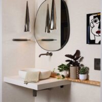 The Modern Mediterranean Powder Room features floating vanity and vessel sink designed to resemble rustic poured concrete; wall mounted faucet in luxe steel finish adding crisp architectural counterpoint. Plaster look wallpaper, burnished steel pendant lights, wall mounted toilet, blackened brass mirror, custom wood ledge, porcelain floor tile designed to mimic terracotta.