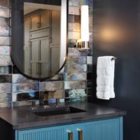 The dark and moody powder room features a custom teal vanity with reeded detailing, and iridescent tile paired with polished French gold hardware; custom legs complete the furniture-like feel, elevating the vanity from functional storage to statement piece. modern yet complementary gold sconces introduce a refined glow to the moody backdrop. Marble asymmetric herringbone floor adds classic pattern and subtle brightness underfoot.