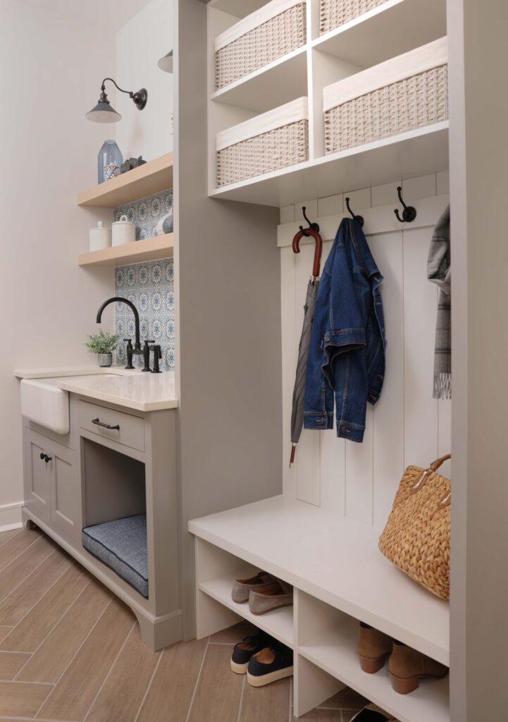 A modern farmhouse mudroom provides plenty of clever storage solutions. Custom cabinetry features a landing area for shoes and jackets, and cubbies to contain smaller items. Above the classic farmhouse sink, rift-cut white oak shelves add natural texture against a striking patterned tile backsplash, while the matte black bridge faucet and fixtures create a clean, modern-edged contrast. A built-in pet nook sits below a durable quartz countertop.