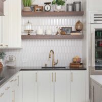 Vintage inspired family kitchen features cherry wood floating shelves, white picket backsplash tile, soft ivory painted cabinets with shaker style doors, black and white quartz countertops, black composite sink, faucet in luxe gold, mixed metal hardware throughout.