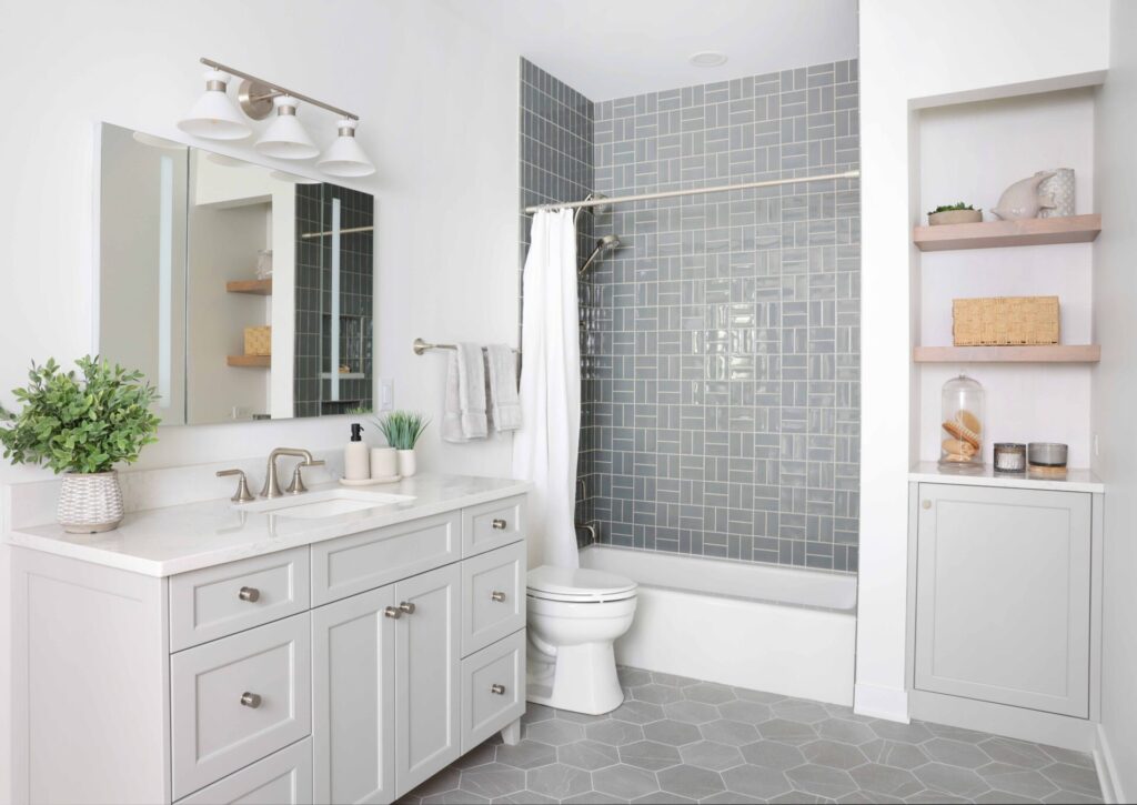 The storage friendly hall bath features a larger vanity with generous drawer storage; a tapered cabinet foot at the base of the vanity gives the piece a furniture-like presence rather than a heavy cabinetry look. Vanity and storage niches are topped with an easy-to-maintain quartz surface selected also for its clean appearance. Finished in a soft gray, the cabinetry keeps the room feeling airy, and brushed nickel hardware brings a luxe element. Also visible is a recessed medicine cabinet with a built-in electrical outlet, a nook with open shelving and additional cabinet storage. In the tub-shower, a gray-blue tile introduces subtle color, and a tile arrangement shift on the back wall adds visual depth. Two shower niches — one lower and one higher — provide easy accessibility to products whether you’re showering or bathing. A hydro rail combines a showerhead and handheld, offering versatility in a cost-effective way.
