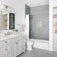 The storage friendly hall bath features a larger vanity with generous drawer storage; a tapered cabinet foot at the base of the vanity gives the piece a furniture-like presence rather than a heavy cabinetry look. Vanity and storage niches are topped with an easy-to-maintain quartz surface selected also for its clean appearance. Finished in a soft gray, the cabinetry keeps the room feeling airy, and brushed nickel hardware brings a luxe element. Also visible is a recessed medicine cabinet with a built-in electrical outlet, a nook with open shelving and additional cabinet storage. In the tub-shower, a gray-blue tile introduces subtle color, and a tile arrangement shift on the back wall adds visual depth. Two shower niches — one lower and one higher — provide easy accessibility to products whether you’re showering or bathing. A hydro rail combines a showerhead and handheld, offering versatility in a cost-effective way.