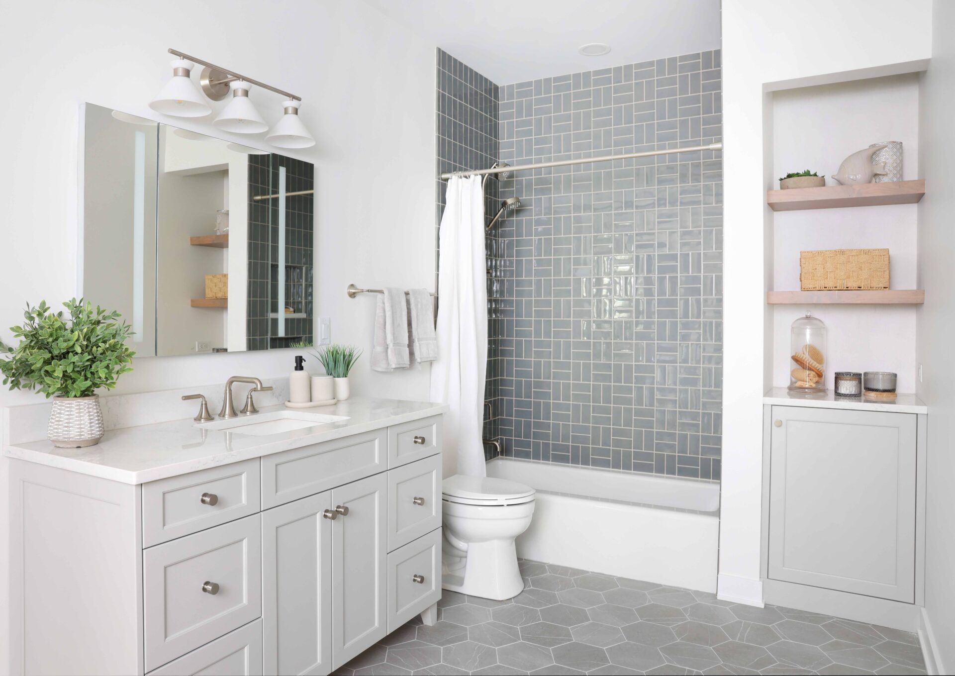 The storage friendly hall bath features a larger vanity with generous drawer storage; a tapered cabinet foot at the base of the vanity gives the piece a furniture-like presence rather than a heavy cabinetry look. Vanity and storage niches are topped with an easy-to-maintain quartz surface selected also for its clean appearance. Finished in a soft gray, the cabinetry keeps the room feeling airy, and brushed nickel hardware brings a luxe element. Also visible is a recessed medicine cabinet with a built-in electrical outlet, a nook with open shelving and additional cabinet storage. In the tub-shower, a gray-blue tile introduces subtle color, and a tile arrangement shift on the back wall adds visual depth. Two shower niches — one lower and one higher — provide easy accessibility to products whether you’re showering or bathing. A hydro rail combines a showerhead and handheld, offering versatility in a cost-effective way.