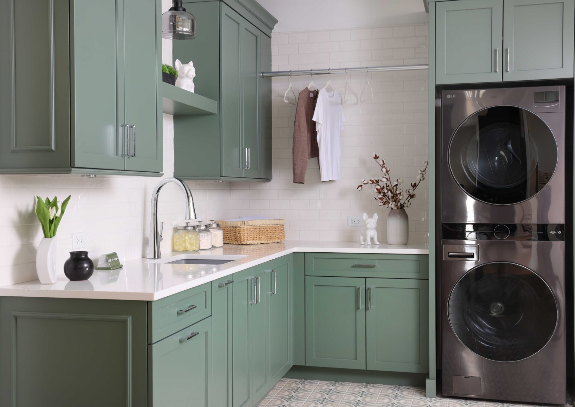 The colorfully classic laundry room features rich green cabinets, floating shelves, subway tile, quartz countertop, gray sink, polished chrome hardware, printed floor tile. Organizational solutions include pull out drying rack, hanging pole for steaming or storing garments, and lots of counter space for folding.