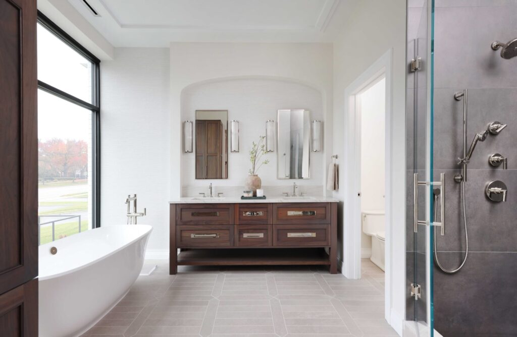 The modern classic primary bath features walnut console vanity topped with quartzite countertop and paired with a textured wallpaper backsplash. Shower contains textured porcelain tile, rain head, shower bench and shaving niche. Freestanding tub makes a sculptural statement on a porcelain tile floor.
