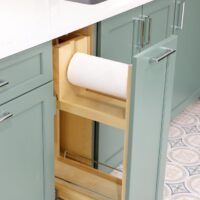Green cabinets in laundry room with pull-out paper towel holder and storage shelf, white quartz countertops and gray sink