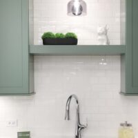 Green painted floating shelf in laundry room with green cabinetry, white subway tile backsplash, polished chrome faucet, white countertops, gray sink, matte black light fixture