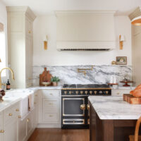 Warm white kitchen with modern vintage details, black and brass statement range, marble countertops and backsplash with shelf, pot filler, red oak island, island seating, farmhouse sink, paneled fridge, wood hood, with sconces, brass hardware and knobs
