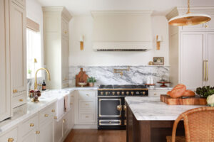 Warm white kitchen with modern vintage details, black and brass statement range, marble countertops and backsplash with shelf, pot filler, red oak island, island seating, farmhouse sink, paneled fridge, wood hood, with sconces, brass hardware and knobs