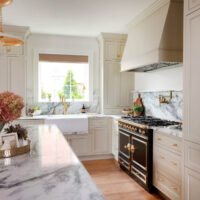 Warm white kitchen with modern vintage details, marble countertops and backsplash with shelf, farmhouse sink with drop ledge, paneled fridge, brass hardware and knobs, to the ceiling cabinets, inset cabinetry, window above sink, set on backsplash in marble, black and brass statement range