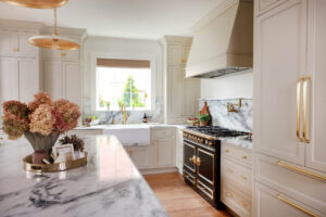 Warm white kitchen with modern vintage details, marble countertops and backsplash with shelf, farmhouse sink with drop ledge, paneled fridge, brass hardware and knobs, to the ceiling cabinets, inset cabinetry, window above sink, set on backsplash in marble, black and brass statement range