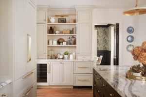 Warm white built-in custom bar with beadboard and brass hardware, paneled refrigerator, to the ceiling cabinets, inset cabinets, crown molding, wine fridge, marble countertops, picture light fixture, brass railing open shelves