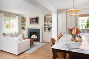 Open concept kitchen and family room, warm white cabinetry, marble countertop on kitchen island, built in shelves on either side of fireplace with picture light fixtures, blue fireplace face
