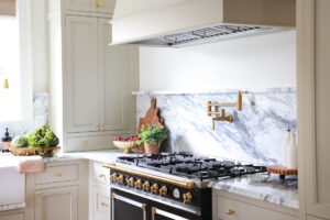 Warm white kitchen with modern vintage details, black and brass statement range, marble countertops and backsplash with shelf, brass pot filler, wood hood, brass hardware and knobs