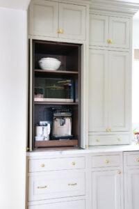 Appliance garage with microwave and coffee machine inside, warm white cabinetry with maple interior. Brass hardware, cabinet latches on top cabinets